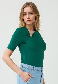 Vrouw met schouderlang haar poseert, gekleed in een groene top met korte mouwen en een ringdetail, gecombineerd met lichtblauwe high-waisted jeans.
