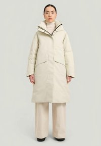 Pufferjacka i ljus beige med hög krage, stora fickor och dragkedja fram. Modellen bär en krämfärgad polo och beige vida byxor.