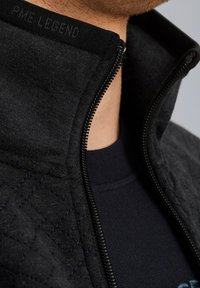 Veste matelassée noire avec une fermeture éclair visible partiellement ouverte, montrant un t-shirt noir en dessous ainsi qu'une partie du cou et du menton d'une personne.