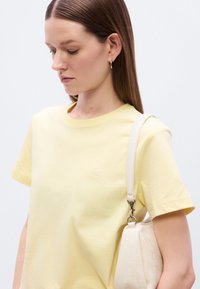 Κίτρινο βαμβακερό μπλουζάκι με στρογγυλή λαιμόκοψη και κοντά μανίκια, με ένα μικρό λευκό κεντημένο λογότυπο. Μια λευκή τσάντα φοριέται στον ώμο.