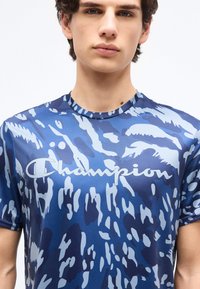 Νεαρός άνδρας που φοράει μπλε μπλουζάκι Champion με σχέδιο καμουφλάζ, εμφανιζόμενος από τους ώμους έως λίγο πάνω από τα χείλη, σε απλό λευκό φόντο.