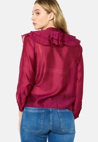 Long-sleeve, maroon blouse featuring a ruffled collar and lightweight, sheer fabric. Pairing with blue denim jeans is visible.