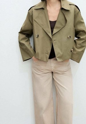 Personne portant une veste courte croisée vert olive, un haut noir et un pantalon beige à jambes larges, les mains dans les poches, sur un fond uni.