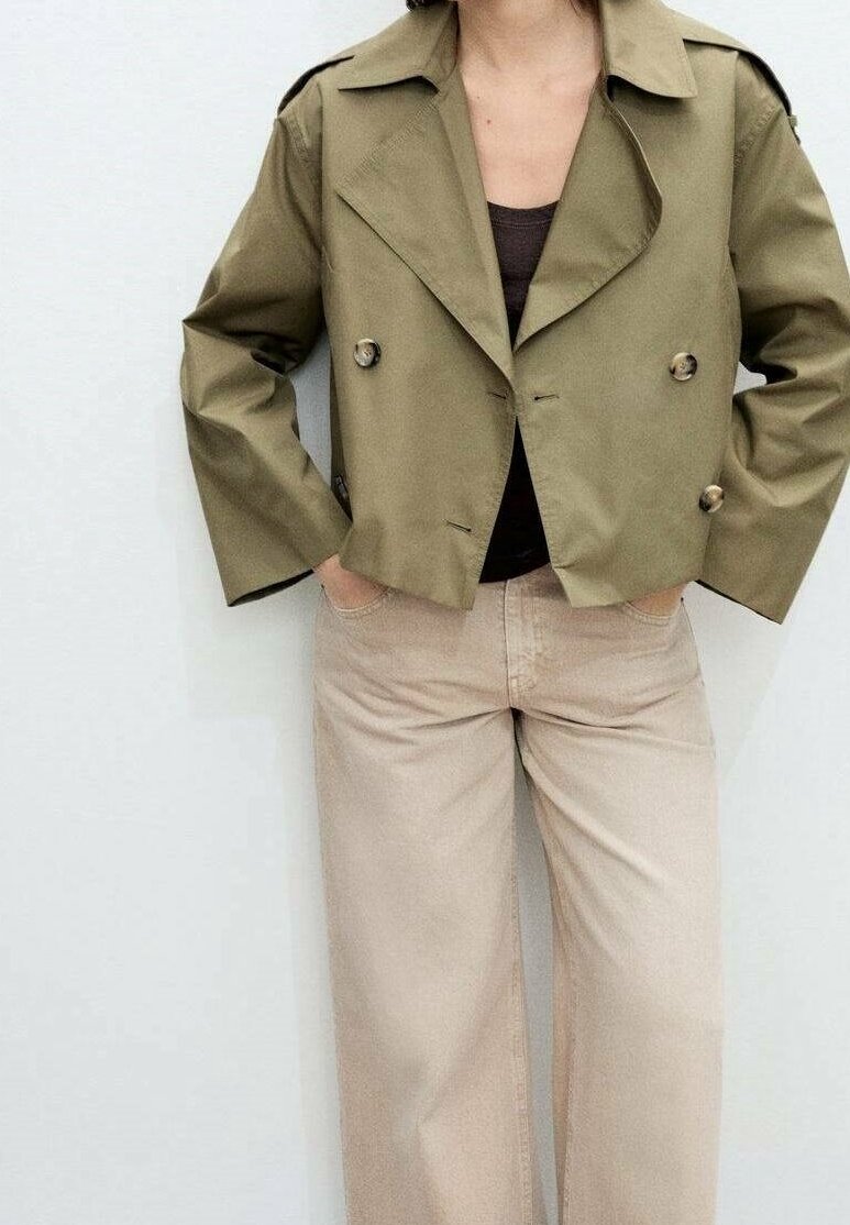 Personne portant une veste courte croisée vert olive, un haut noir et un pantalon beige à jambes larges, les mains dans les poches, sur un fond uni.