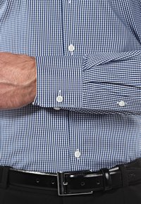 Camicia a scacchi blu e bianca con polsini abbottonati, realizzata in tessuto leggero. Presenta un motivo a gingham stretto e una cintura nera.