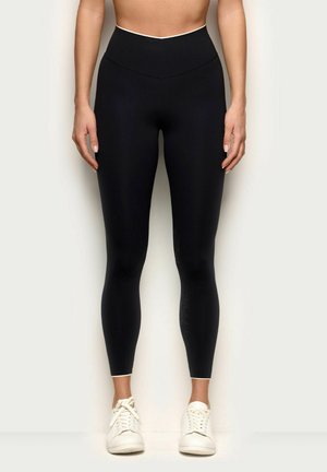 Mujer con leggins negros de talle alto y zapatillas blancas, de pie contra un fondo claro y liso con los brazos relajados a los lados.