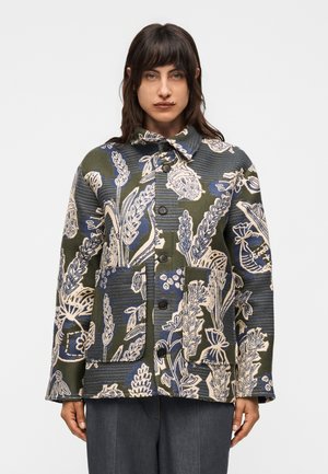 Γυναίκα με πράσινο, μπλε και μπεζ μπουφάν με κουμπιά και λουλουδάτο μοτίβο, με δύο μπροστινές τσέπες, στέκεται μπροστά σε απλό λευκό φόντο.