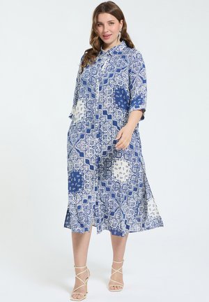 Frau in blau-weiß gemustertem Midikleid mit Kragen und 3/4-Ärmeln, trägt beige Riemchensandaletten mit hohen Absätzen.