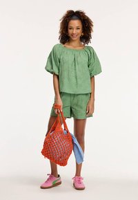 Zeleni dvodijelni outfit s teksturom koji uključuje gornji dio s naduvanim rukavima i odgovarajuće kratke hlače. Dodaci su velika narančasta mrežasta torba i ružičaste tenisice.