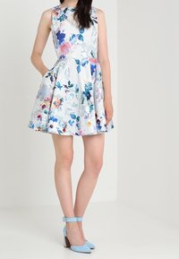 Robe sans manches à imprimé floral en blanc avec des accents bleus, roses et verts ; jupe évasée. Associée à des escarpins à bride cheville bleu clair.