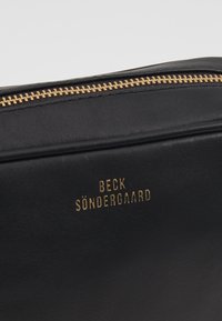 Zbliżenie czarnej skórzanej torby z złotym zamkiem błyskawicznym i złotym wytłoczonym napisem "Beck Söndergaard" z przodu.