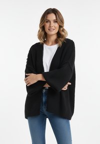 Czarny dziergany kardigan o szerokich rękawach, z otwartym frontem, wykonany z teksturowanej tkaniny, noszony na białym topie, w połączeniu z niebieskimi dżinsami.