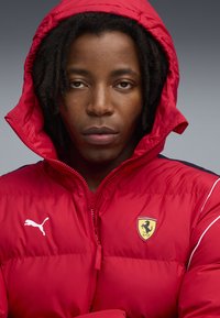 Rød puffjakke med hætte, med hvidt Puma-logo på brystet og et gult Ferrari-emblem. Glat, skinnende overflade og sporty design.