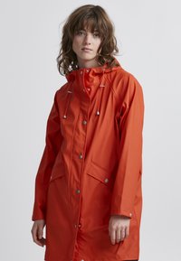 Cappotto impermeabile arancione con cappuccio, chiusure a scatto, tasche laterali e coulisse regolabile. Tessuto liscio con una vestibilità elegante e leggermente ampia.