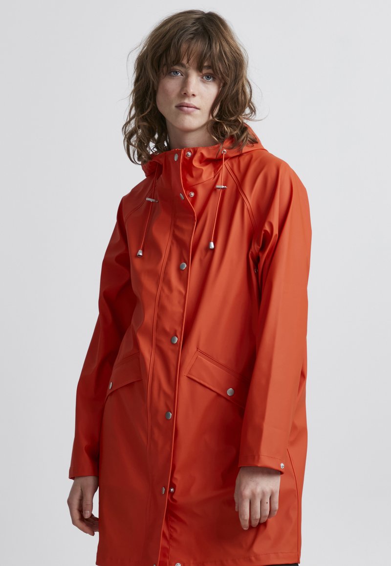 Cappotto impermeabile arancione con cappuccio, chiusure a scatto, tasche laterali e coulisse regolabile. Tessuto liscio con una vestibilità elegante e leggermente ampia.