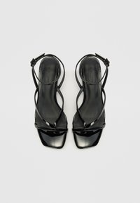 Sandales en cuir verni noir avec des brides croisées élaborées, bouts carrés et brides de cheville réglables. Texture lisse avec finition brillante.