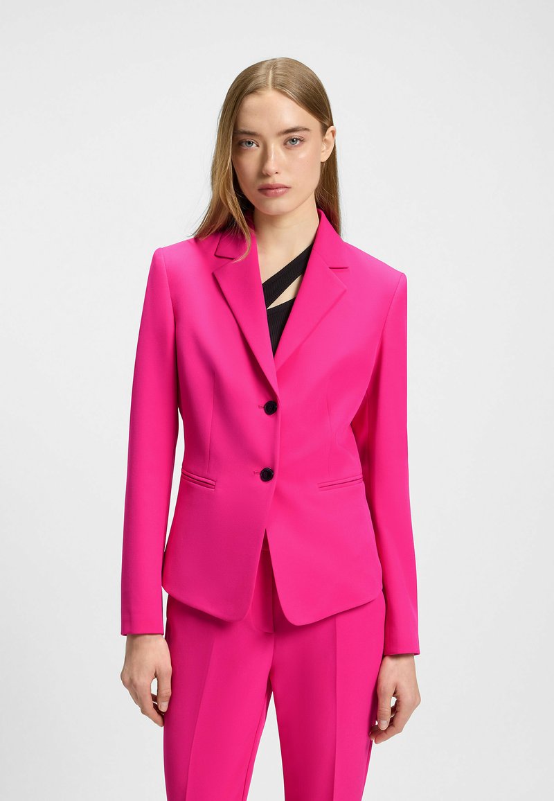 Blazer fuchsia à deux boutons, avec des revers crantés et des poches à rabat à l'avant. Présente une texture lisse et une coupe structurée, associé à un pantalon assorti.