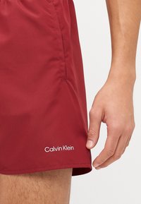 Shorts de bain bordeaux avec une texture lisse, arborant un logo blanc "Calvin Klein" près de l'ourlet et un détail de poche sur le côté.