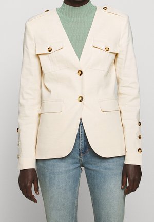 Krémový lněný blazer s dvojitými náprsními kapsami, zlatými knoflíky a přiléhavým střihem. Nosený přes strukturovaný zelený top a modré džíny.