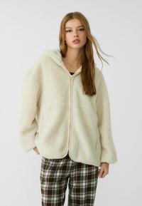 Cremefarbene, mit Fleece gefütterte Zip-Jacke mit Kapuze, die einen lässigen Schnitt und zwei Seitentaschen aufweist. Getragen über karierter brauner und beiger Hose.