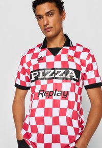 Rød og hvid ternet trøje med en sort krave og logoer. Forsiden viser "PIZZA" og "Replay" i fed hvid tekst.