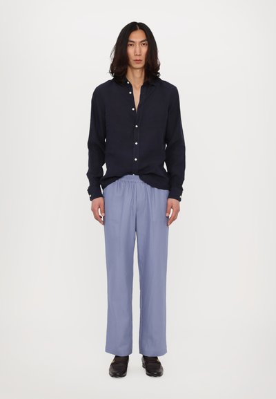 Cămașă albastră cu mânecă lungă și nasturi, combinată cu pantaloni elastici de culoare albastră deschisă și pantofi negri, stând în fața unui fundal alb.