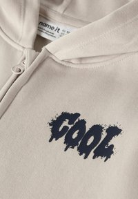 Beige hettegenser med glidelås og teksturert marineblå «COOL»-trykk på forsiden. Mykt stoff, standard hette og synlige sømdetaljer.