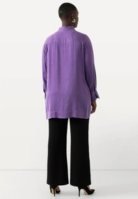 Camicia viola a maniche lunghe con vestibilità rilassata, dettaglio con piega sul retro e polsini abbottonati, abbinata a pantaloni neri a gamba larga e tacchi alti.