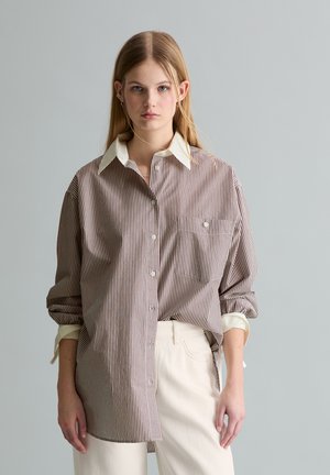 Młoda kobieta ubrana w dużą paskowaną koszulę na guziki z podwiniętymi rękawami i kremowe spodnie na jednolitym szarym tle.