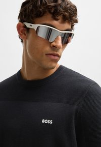 Naočale s bijelim okvirom i zrcalnim staklima, nosene uz crni pulover s teksturom koji ima horizontalni rebrasti uzorak i "BOSS" logotip.