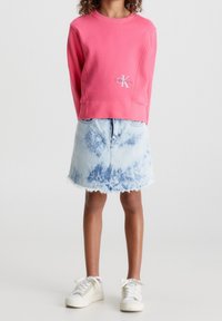 Felpa rosa con logo Calvin Klein, abbinata a una gonna di jeans blu chiaro con orlo sfrangiato. Sneakers bianche completano l'outfit.