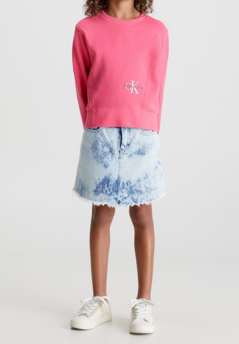 Felpa rosa con logo Calvin Klein, abbinata a una gonna di jeans blu chiaro con orlo sfrangiato. Sneakers bianche completano l'outfit.