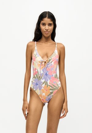 Femme aux longs cheveux foncés portant un maillot de bain une pièce à fleurs avec un détail de nœud à l'avant sur un fond clair uni.