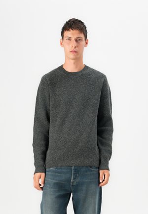 Grijze gebreide trui met een ronde hals en geribbelde textuur, gecombineerd met lichtblauwe jeans. De stof lijkt zacht en warm.