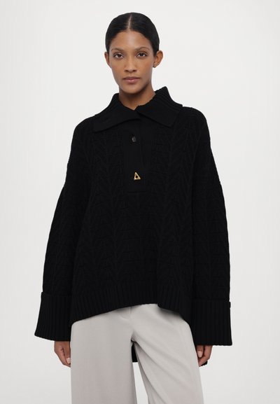 Black knit pullover sweater with a prominent collar, textured pattern, button details, and ribbed cuffs, paired with light gray pants.