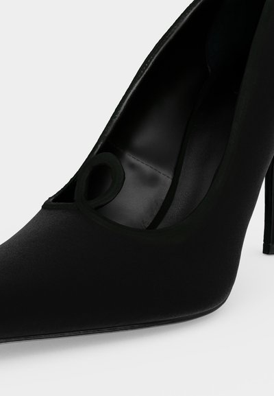 Chaussure noire à talon haut avec un bout pointu, une finition lisse et une petite boucle décorative près de l’ouverture.