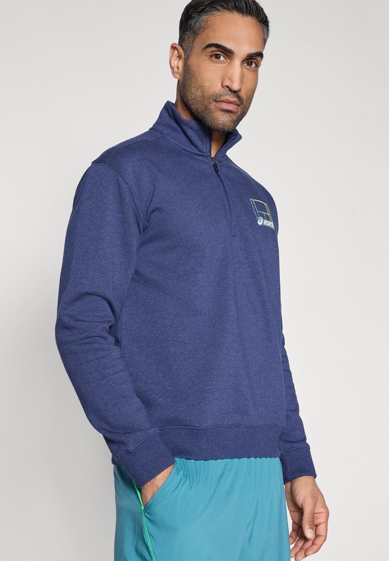 Sweat-shirt à fermeture éclair bleu marine en tissu doux et texturé avec un ourlet et des poignets côtelés. Présente un logo vert clair sur la poitrine.