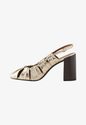 Pied portant des chaussures à talons hauts métalliques dorées avec des détails découpés, posé sur un sol gris lisse.