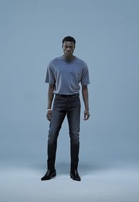 T-shirt cinzenta de mangas curtas combinada com jeans de ganga escura. Modelo usa sapatos pretos e uma pulseira prateada, posando contra um fundo azul claro.