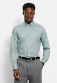 Hellgrünes Hemd mit Muster, spitzem Kragen, Knopfleiste und langen Ärmeln, kombiniert mit grauer Hose und schwarzem Gürtel.