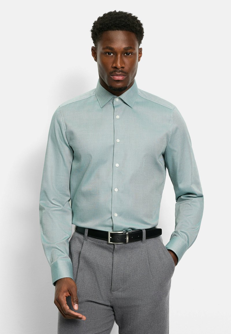 Hellgrünes Hemd mit Muster, spitzem Kragen, Knopfleiste und langen Ärmeln, kombiniert mit grauer Hose und schwarzem Gürtel.