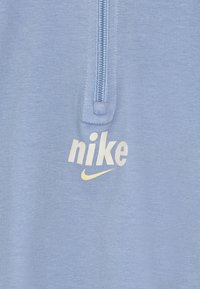 Svijetloplavi dukserica s četvrtim patentnim zatvaračem napravljena od mekog materijala, s bijelim "Nike" logotipom i žutim swooshom sa prednje strane. Teksturalna površina.