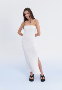 Weißes schnittloses Kleid aus strukturiertem Stoff, Midi-Länge und seitlichem Schlitz. Getragen mit schwarzen Plateausandalen. Einfaches und figurbetontes Design.