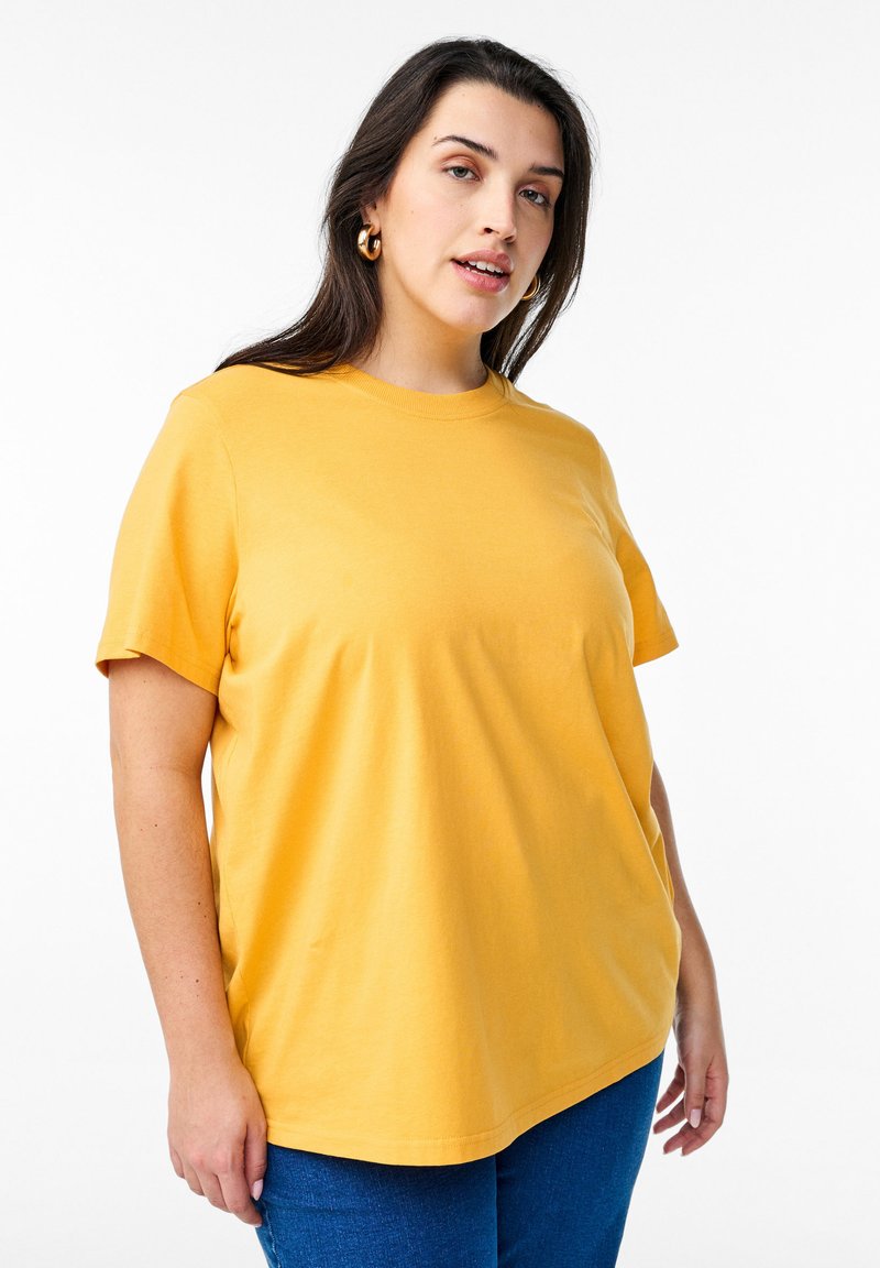 Femme aux longs cheveux brun foncé portant une chemise jaune à manches courtes et un jean bleu, se tenant devant un fond blanc avec une expression neutre.