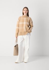 Sweater de malha xadrez bege com gola alta, combinado com calças largas brancas. Modelo segura uma bolsa tote bege. Textura suave e corte descontraído.