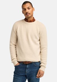 Maglione beige a coste con maniche raglan, colletto rotondo e finituraTesturizzata, indossato sopra una camicia a quadri, abbinato a jeans blu scuro.