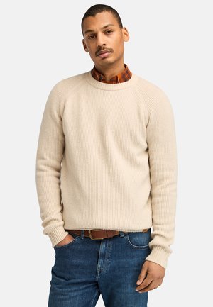 Maglione beige a coste con maniche raglan, colletto rotondo e finituraTesturizzata, indossato sopra una camicia a quadri, abbinato a jeans blu scuro.