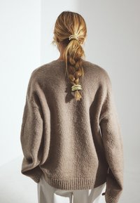 Tekstūrēts brūns pārmēru liels krekls ar plašām piedurknēm un ribināta apakšmalu, skatīts no aizmugures. Mati iekārtoti zirgaste, nostiprināti ar zelta spraudni.