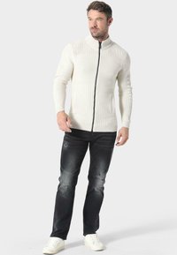 Suéter blanco de punto con cremallera, textura acanalada, cuello alto y bolsillos laterales, combinado con jeans oscuros desgastados y zapatillas blancas.