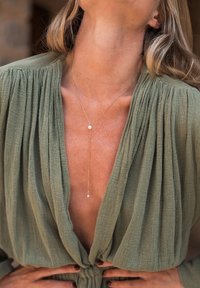 Gold pendant necklace with a fine chain featuring a single pearl accent, worn over a deep v-neck olive green textured fabric top.